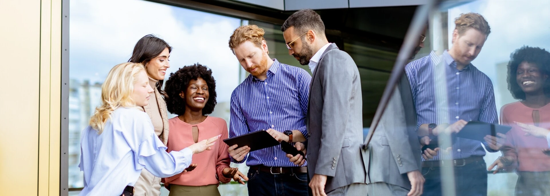 Eine diverse Gruppe von Geschäftsleuten diskutiert gemeinsam über ein Tablet vor einem Bürogebäude.