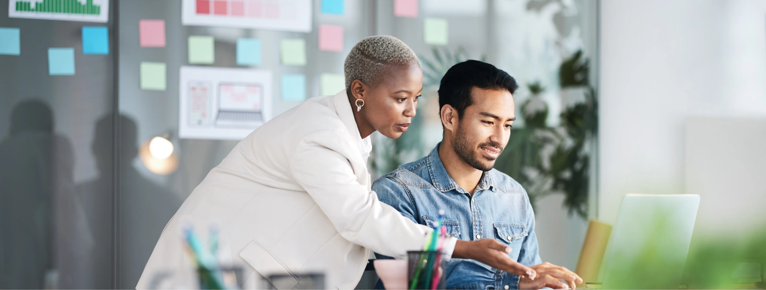 Eine Frau und ein Mann arbeiten zusammen an einem Laptop im Büro. Die Frau erklärt dem Mann etwas.