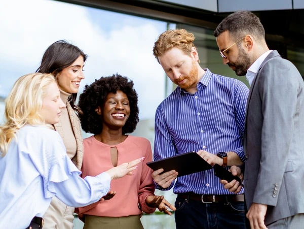 Eine diverse Gruppe von Geschäftsleuten diskutiert gemeinsam über ein Tablet vor einem Bürogebäude.
